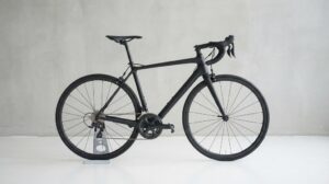 薄いグレーのコンクリート壁の前で、無地のマットな黒いロードバイクが小さなスタンドに立てられ、真横から写された商品写真のようなカット