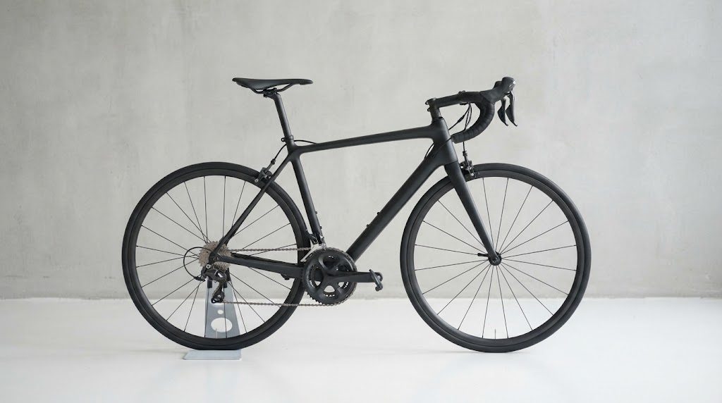 薄いグレーのコンクリート壁の前で、無地のマットな黒いロードバイクが小さなスタンドに立てられ、真横から写された商品写真のようなカット