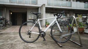曇りの朝、集合住宅の玄関前の駐輪スペースに、無地の白いロードバイクがラックの横に停められ、背景に植木鉢と他の自転車が写る様子