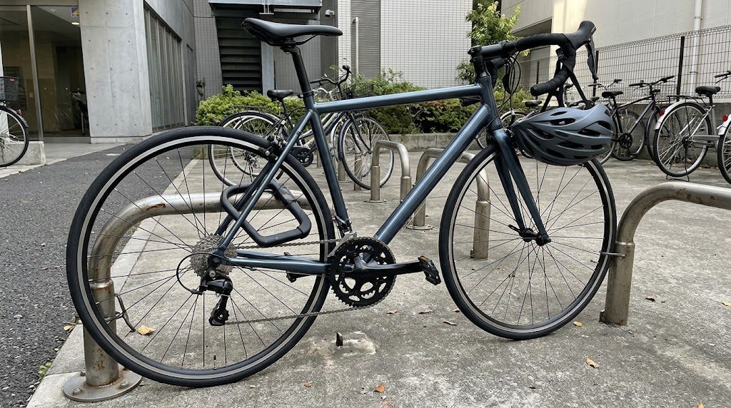 オフィスの駐輪場で、ロードバイクがフレームと後輪をU字ロックでラックに固定されて停められ、ハンドルにヘルメットが掛けられている様子