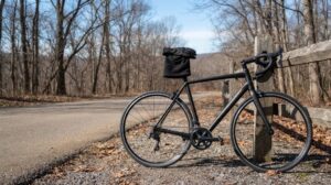 冬の乾いた道の脇に立てかけられたロードバイクのサドルに、冬用の手袋とネックウォーマーが掛けられている様子。背景は葉の落ちた木々