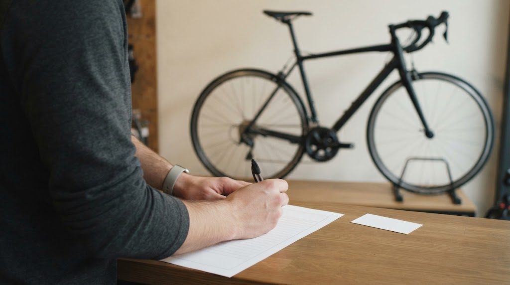 自転車店のカウンターで、罫線だけの白い用紙に片手でペンを持って書き込む手元。奥にロードバイクが見える